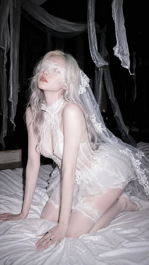 A pale-haired woman in a white lace outfit kneels on a bed beneath hanging fabric in a dark interior.